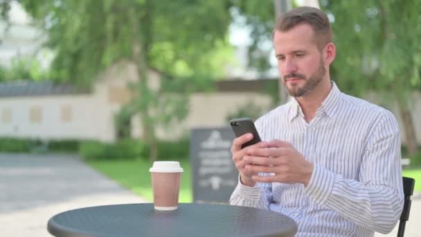 Hombre de mediana edad celebrando el éxito en el teléfono inteligente en el café al aire libre — Vídeos de Stock