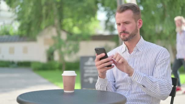 Medelålders man reagerar på förlust på Smartphone i utomhus Cafe — Stockvideo