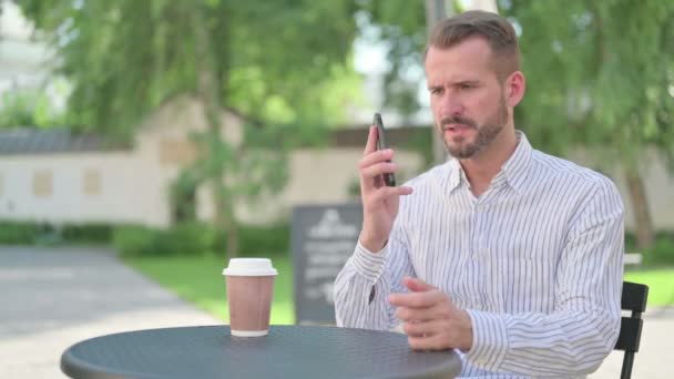 Hombre de mediana edad enojado hablando en el teléfono inteligente en el café al aire libre — Vídeos de Stock