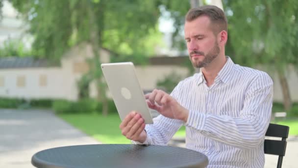 Medelålders man reagerar på förlust på surfplatta i utomhus Cafe — Stockvideo