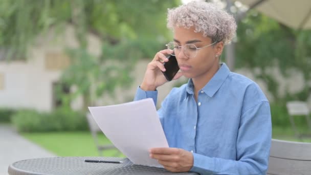 Joven mujer africana hablando por teléfono, leyendo documentos en el café al aire libre — Vídeos de Stock