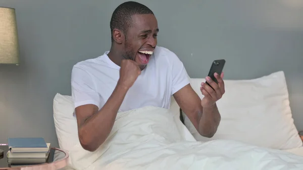 Sucesso, Jovem Africano Celebrando no Smartphone na cama — Fotografia de Stock