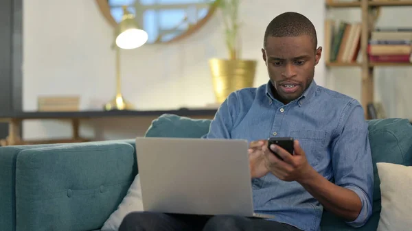 Junger Afrikaner arbeitet mit Smartphone und Laptop auf Sofa — Stockfoto