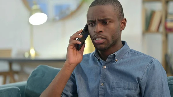 Joven hombre africano enojado en el teléfono inteligente en el sofá —  Fotos de Stock