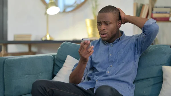 Hombre joven africano reaccionando a la pérdida en el teléfono inteligente en el sofá —  Fotos de Stock