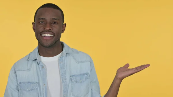 Young African Man Holding Product on Palm, Yellow Background — Stock Photo, Image