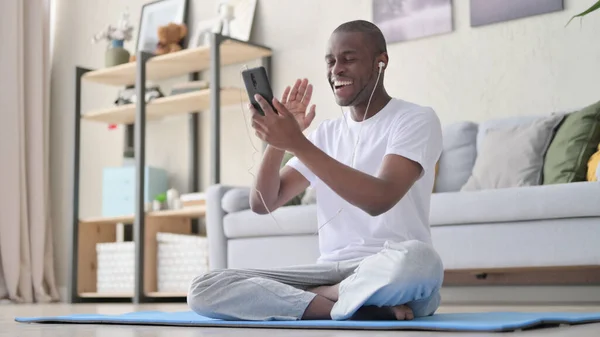 Hombre africano hablando en videollamada mientras está sentado en la esterilla de yoga —  Fotos de Stock