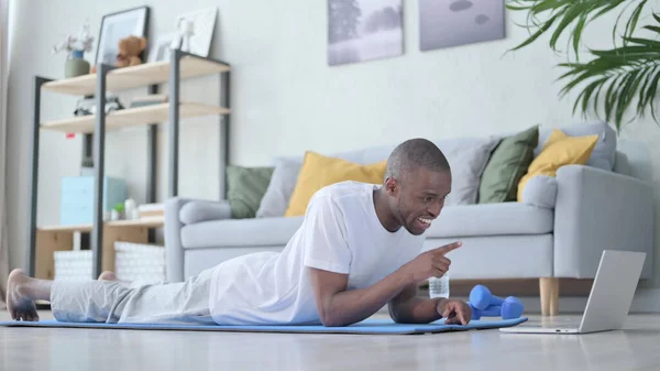 Hombre africano haciendo videollamada en el portátil mientras está acostado en la esterilla de yoga —  Fotos de Stock