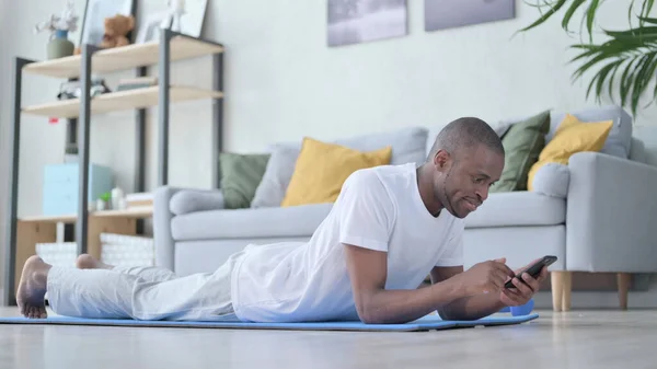 Afrikai férfi okostelefonnal feküdt a jóga matracon — Stock Fotó