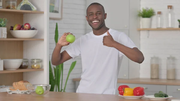 African Man Showing Thumbs up while Holding Apple in Kitchen — ストック写真