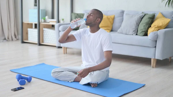 Hombre africano bebiendo agua en la esterilla de yoga en casa — Foto de Stock