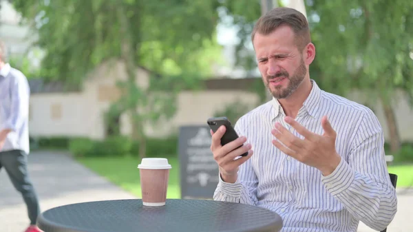 Hombre adulto maduro molesto por la pérdida en el teléfono inteligente en el café al aire libre — Foto de Stock