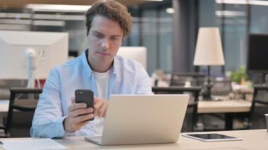 Ofiste dizüstü bilgisayar kullanırken akıllı telefon kullanan adam 