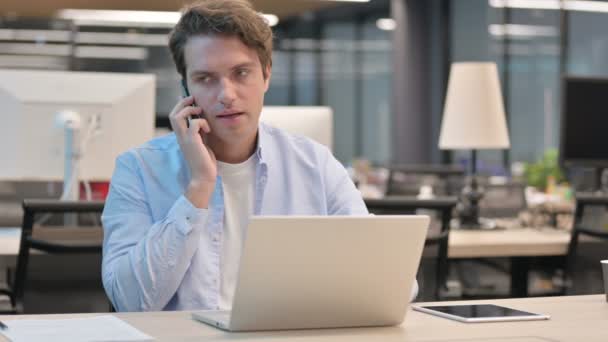 Mann spricht im Büro mit Laptop auf Smartphone — Stockvideo