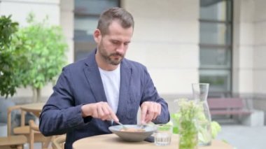 Adam otururken yemek yiyor, açık kafede oturuyor. 