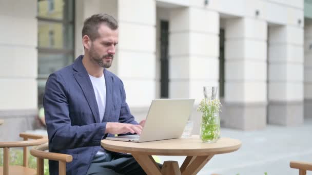 Homme ayant mal au dos tout en utilisant un ordinateur portable, assis dans un café en plein air — Video