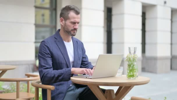 Muž s bolestí hlavy pracuje na notebooku, sedí v venkovní kavárně — Stock video
