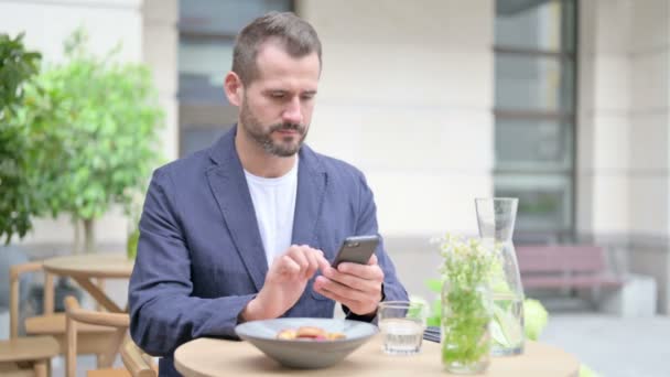 Man Browsing Internet op Smartphone, Zittend in Outdoor Cafe, Voorkant Pose — Stockvideo