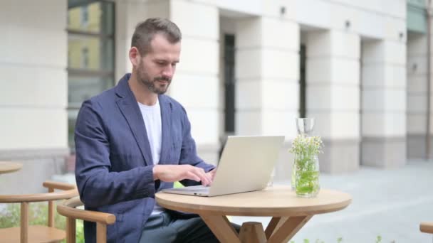 Uomo che punta alla fotocamera durante l'utilizzo del computer portatile, seduto in caffè all'aperto — Video Stock