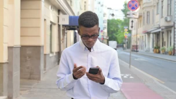 African Man reaguje na straty na smartfonie podczas spaceru po ulicy — Wideo stockowe