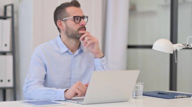 Orta yaşlı adam iş yerinde dizüstü bilgisayar kullanırken düşünüyor 