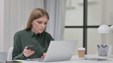 Laptop üzerinde çalışırken akıllı telefon kullanan genç kadın.