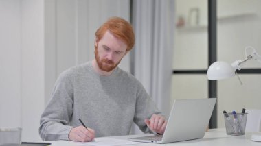 Kızıl Sakallı Adam Kağıda Yazarken Dizüstü bilgisayar kullanıyor 