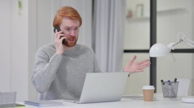 Dizüstü bilgisayarlı kızgın sakallı kızıl saçlı adam akıllı telefondan konuşuyor. 