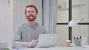 Dizüstü bilgisayarlı genç sakallı kızıl adam Kameraya gülümsüyor 