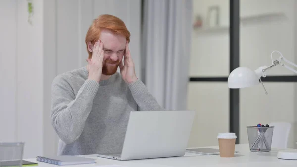 Homem ruivo de barba com dor de cabeça enquanto digita no laptop — Fotografia de Stock