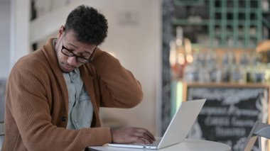 Kafede dizüstü bilgisayar kullanırken boynu ağrıyan Afrikalı bir adam. 
