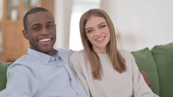 Casal Interracial sentado no sofá e sorrindo para a câmera — Fotografia de Stock