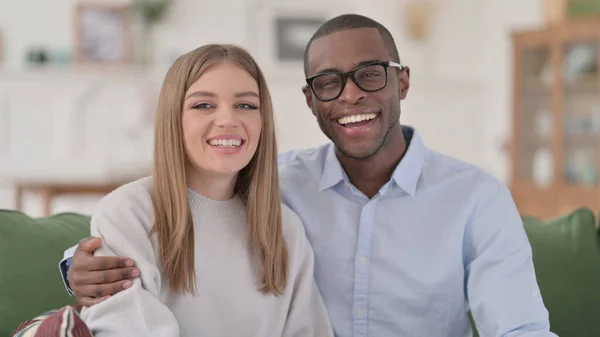 Abraçando o casal inter-racial Sorrindo para a câmera enquanto sentado no sofá — Fotografia de Stock