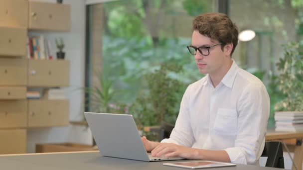 Hombre joven mirando a la cámara mientras usa el ordenador portátil en la oficina moderna — Vídeos de Stock