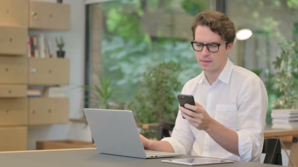 Homem irritado falando em Smartphone ao usar laptop no escritório moderno — Vídeo de Stock