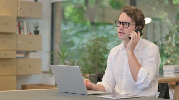 Hombre joven hablando en Smartphone mientras usa el ordenador portátil en la oficina moderna — Vídeos de Stock