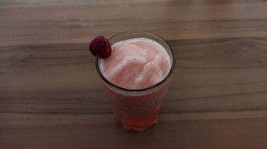 Ahşap bir masada uzun bir bardakta ahududu süslemeli ferahlatıcı pembe meyve püresi. Parlak doğal ışık. Canlı ve davetkar bir atmosfer yaratıyor..