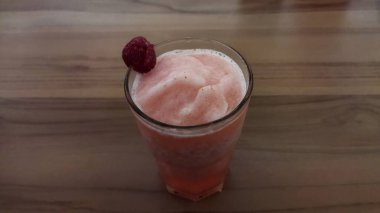 Üzerinde ahududu süslemesi olan, ahşap bir masada oturan, parlak ve davetkar bir atmosfer yaratan, sağlık ve içecek pazarlaması için mükemmel bir ahududu içeren ferahlatıcı pembe meyve püresi..