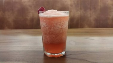 Buzlu, köpüklü pembe meyve kokteyli ve ahudududulu garnitür ahşap bir masada berrak bir bardakta ferahlatıcı ve davetkar bir atmosfer yaratır içecek pazarlama ve konukseverlik promosyonu için mükemmel..