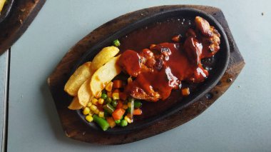 Koyu oval bir tabakta barbekü sosuyla kaplanmış sulu et yanında kızarmış patates kızartması ve doğal ışıklandırmayla karışık renkli sebzeler..