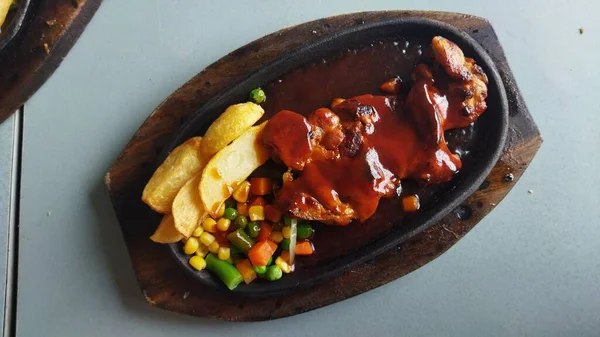 Koyu oval bir tabakta barbekü sosuyla kaplanmış sulu et yanında kızarmış patates kızartması ve doğal ışıklandırmayla karışık renkli sebzeler..