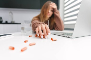 Mutfakta dizüstü bilgisayarla oturan kadın kırmızı haplar alıyor.