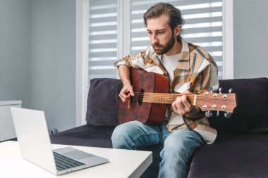Evden çalışıyorum. Genç bir adam gitar çalmayı öğreniyor. Online dersler. E-öğrenme. Uzaklık öğrenimi.