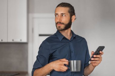 Young man at home holding a mobile phone and a mug and looking to the side