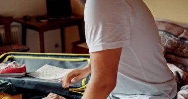 Adam bavulunu topluyor, yatakta oturuyor. Sınırlar açıldıktan sonra yaz tatilinde seyahat için bavulları hazırlamak. Adam gülümsüyor, yaklaşan gezi için heyecanlı. Yol gezisi kavramı, kısıtlamaların kaldırılması.