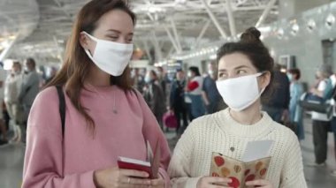 Koruyucu maskeli insanlar uçağa binmek için sırada bekliyorlar. Tıbbi maskeli iki kadın havaalanı terminalindeki kameraya bakıp kendilerini koronavirüsten uzak tutuyorlar. Salgın covid konsepti
