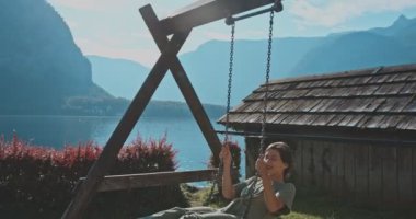 Salıncakta sallanan kaygısız genç bir kadın arka planda muhteşem bir doğa ile harika vakit geçiriyor. Kız anın tadını çıkarıyor ve ağaçlı ve suyla dağların görkemli manzarasına hayran. Seyahat kavramı, güzellik.