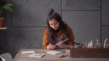 Önlüklü bir kadın çömlekçi, tezgahın üstüne nazikçe bir parça kilden yoğurarak elleriyle çalışıyor. Arkadaki raflarda çömlek ve pencereden güneş ışığı. Küçük bir iş kurun.