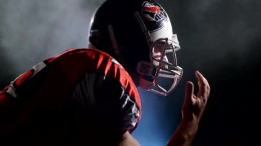 Profesyonel takım sporu kavramı. Parlak stadyum ışığında gece maçına hazırlanan kararlı Amerikan futbolcusunun silueti. Üniformalı ve kasklı sporcu saldırıya hazır, izole edilmiş..