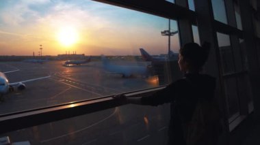 Genç bayan gezgin, boş zaman ya da iş yeri büyük uluslararası havaalanı merkezinin cam penceresinin yanında duruyor, gündoğumu arka planı. Boş havaalanı terminali kapalı salon, yalnız bayan yolcu bekliyor.
